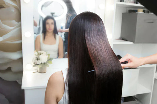 A stylist carefully combs long, shiny hair in front of a well-lit mirror, with a bouquet of flowers on the vanity.