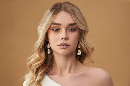 A woman with wavy golden blonde hair and pearl earrings gazes confidently at the camera. She wears an off-shoulder white top against a soft brown background.