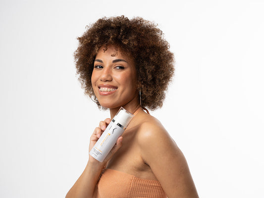A woman with curly hair smiles while holding a GK Hair Leave-In Conditioner against her shoulder. She wears a strapless top, conveying confidence and beauty.