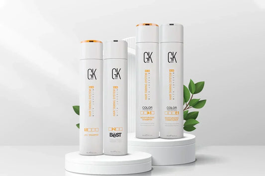 Four sleek hair care products from GK Hair, including a shampoo and conditioner, displayed on white pedestals with green leaves.