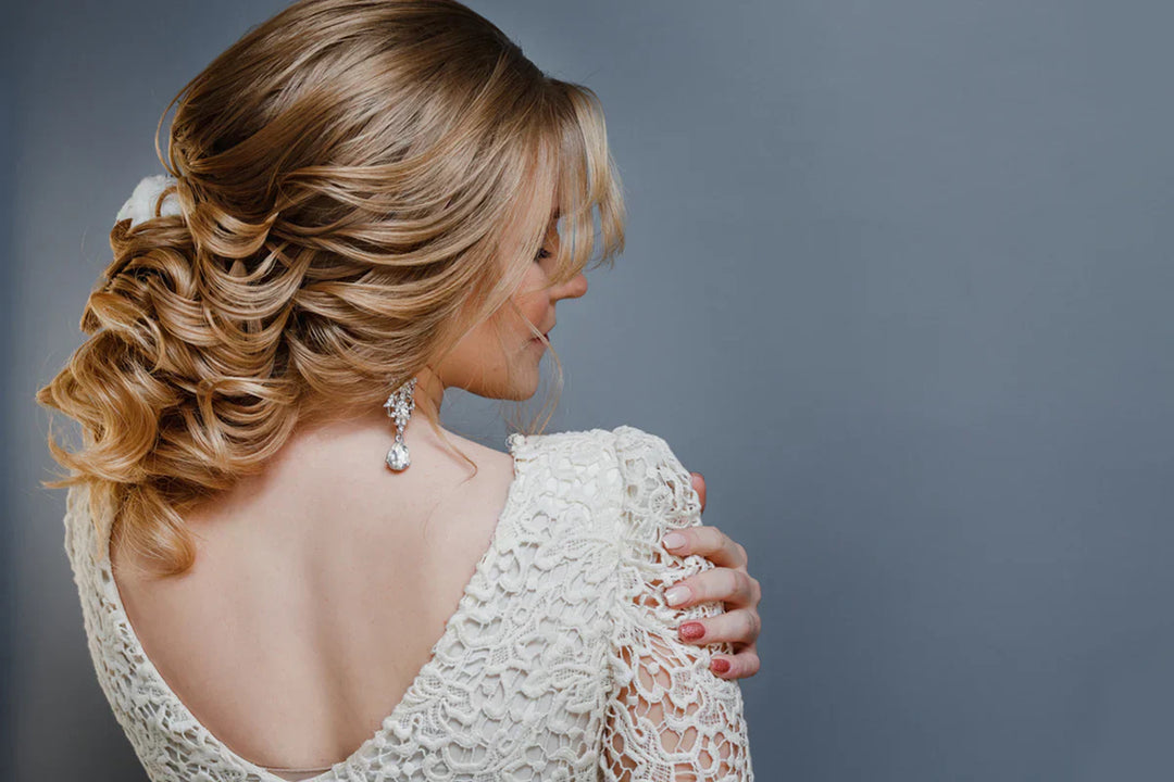 A woman with wavy gold blonde hair styled in an elegant updo, wearing a lace white dress and pearl earrings, gazes downwards, exuding a serene mood.