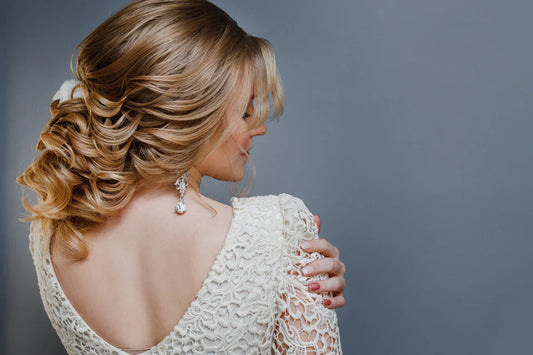 A woman with wavy gold blonde hair styled in an elegant updo, wearing a lace white dress and pearl earrings, gazes downwards, exuding a serene mood.