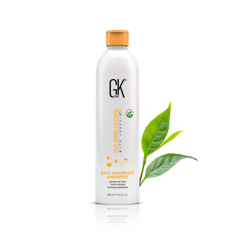 GK Hair Anti-Dandruff Shampoo 250 ml white bottle with orange cap beside two green tea leaves, reflected on white